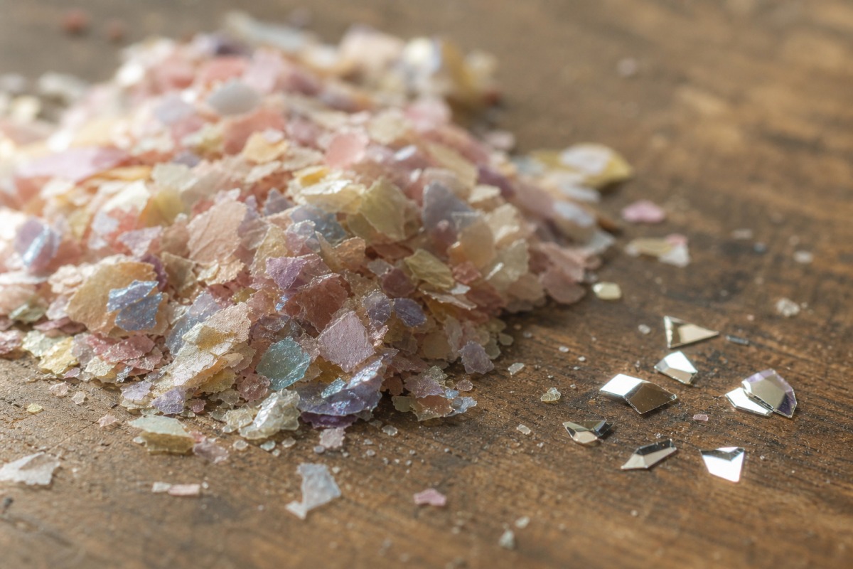 Close-up of a small heap of more matte cellulose bio-glitter beside a few shiny synthetic PET particles on a wooden surface.