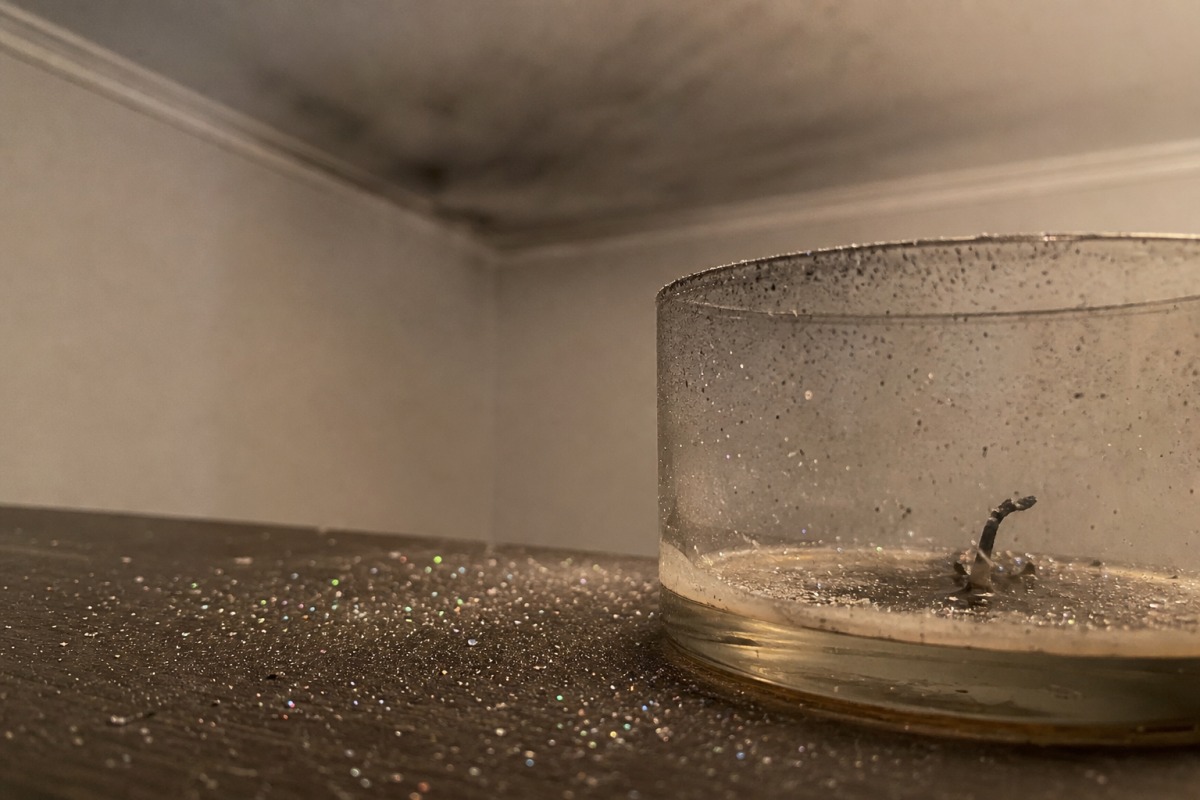 Tiny glitter particles scattered across a table around a burned-out candle, with a thin grey film of soot on the ceiling in the background.