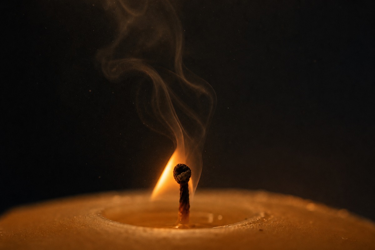 A candle wick with an unstable flame and a rising trail of black smoke in a dark room